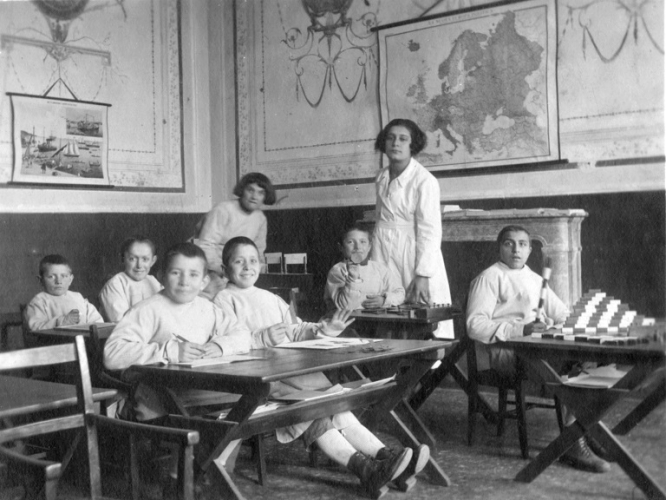 Aula scolastica della Colonia-Scuola “Antonio Marro” di Reggio Emilia con alunni ai banchi e una maestra in piedi, dicembre 1921 (Archivio del San Lazzaro, Reggio Emilia)